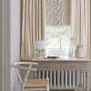 Cream pleated curtains framing a patterned Roman shade with branch motif; shade partially lowered, filtering soft daylight onto a pale wooden desk with lamp, chair, vases beside a garden view.