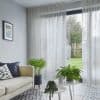 Sheer grey curtains on a ceiling track, drawn across a sliding glass door and filtering daylight while slightly parted to show a lawn; living room with sofa, patterned tile and plants.
