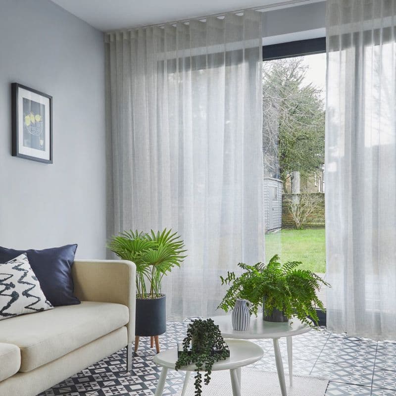 Sheer grey curtains on a ceiling track, drawn across a sliding glass door and filtering daylight while slightly parted to show a lawn; living room with sofa, patterned tile and plants.