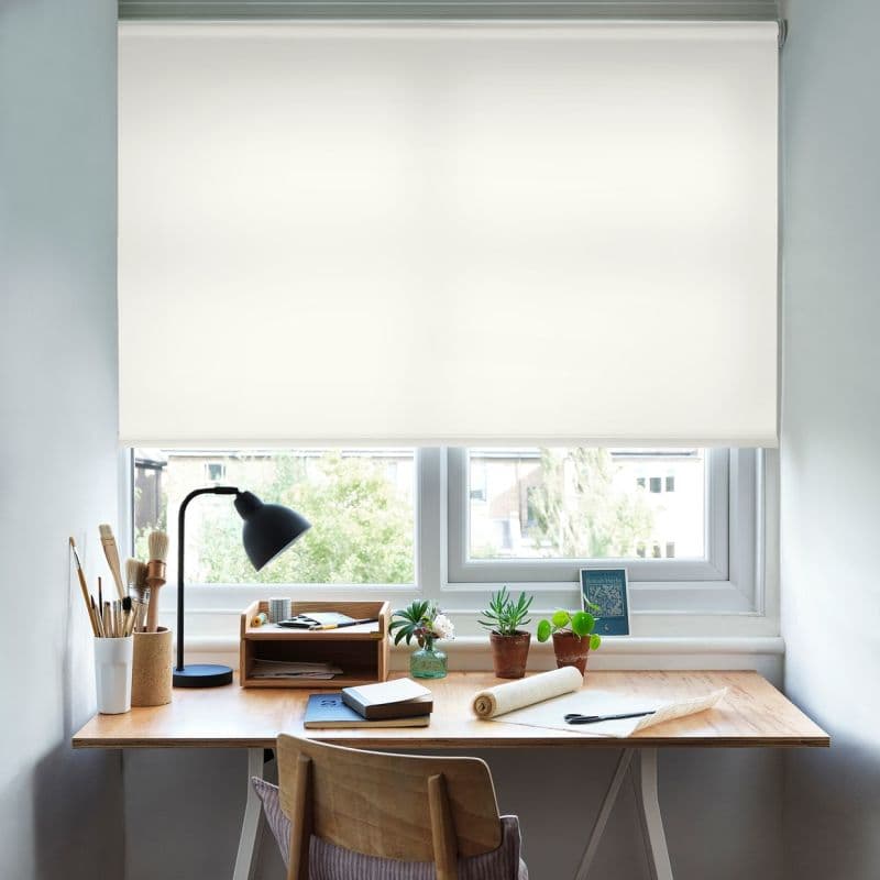 White fabric roller blind fully lowered, diffusing bright daylight; a wooden desk beneath holds a lamp, brushes, notebooks and potted plants — a study with natural light and view.