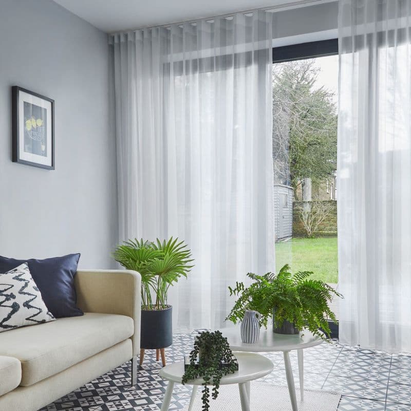 Sheer white floor-length pleated curtains on a ceiling track, filtering daylight and slightly parted to reveal a grassy backyard; modern living room with beige sofa, patterned tile floor and plants.