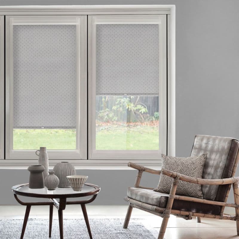 Patterned gray roller blinds covering a three-panel window—left fully lowered, right half-lowered—filtering daylight. Neutral living room with a rattan armchair, side table and a soft garden view outside.