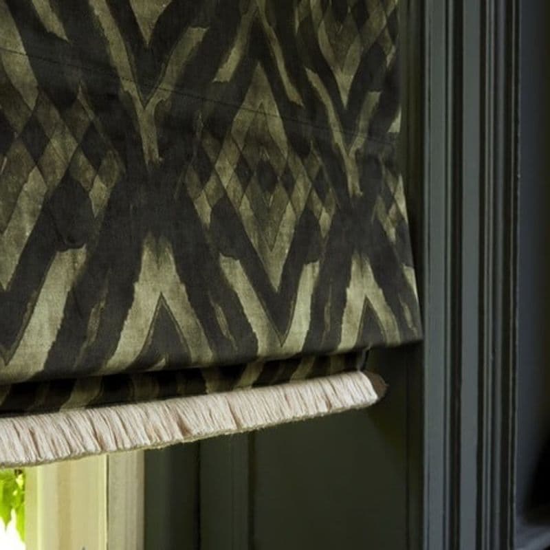 A patterned fabric Roman shade with olive-and-black geometric/ikat design and pale fringe trim hangs partially lowered and folded, covering a window within dark green trim, softly lit from the left.