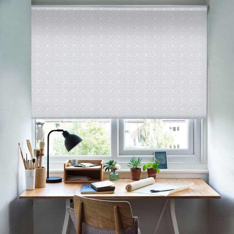 A patterned white roller blind, fully lowered across a double window, diffusing daylight; a wooden desk beneath with lamp, art supplies, potted succulents and a chair in a bright workspace.