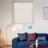 Beige pleated cellular blind, lowered over the window with horizontal texture, blocking most light while a narrow bottom gap reveals a rooftop; in a bright living room with blue sofa, pillows, lamp.