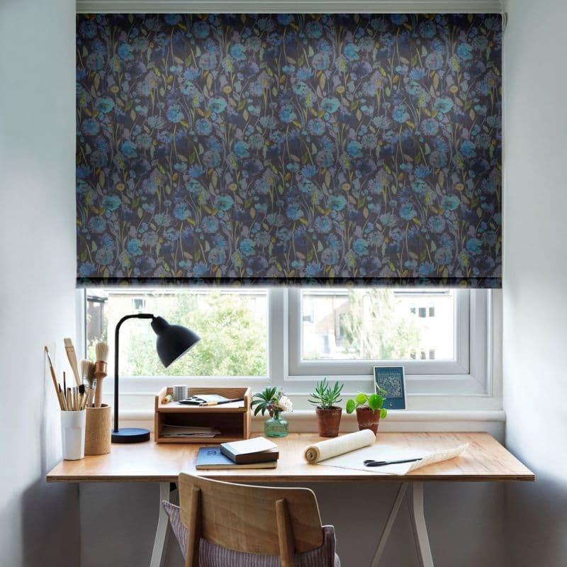 Floral roller blind with dark background and blue-green flowers, fully lowered over window, filtering light; a home desk beneath holds a lamp, paintbrushes, potted succulents and a wooden chair.