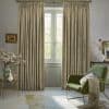 Beige pleated patterned curtains on a brass rod, drawn to frame a central glass French door, filtering soft daylight into a living room with a green armchair and decorative vases.