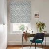 Blue-and-white floral Roman blind lowered over window, partially filtering daylight; desk with teal chair, small mirror and vase of flowers in a softly lit neutral study with framed seaside print.