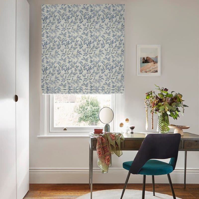 Blue-and-white floral Roman blind lowered over window, partially filtering daylight; desk with teal chair, small mirror and vase of flowers in a softly lit neutral study with framed seaside print.