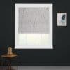 Patterned Roman blind lowered, filtering soft daylight through a white-framed window on a dark-gray wall; wooden side table with glass-dome lamp left, abstract art right, and blurred greenery beyond.