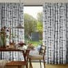 Floor-to-ceiling pleated curtains with a vertical black-blue watercolor brushstroke pattern, parted centrally to reveal a sunlit backyard; framing a dining table set for breakfast with wooden chairs and pendant lamp.