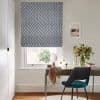 Blue-grey chevron Roman blind lowered over a sash window, filtering soft daylight; home office desk beneath holds a mirror, scarf, vase of flowers and teal-upholstered chair against neutral walls.