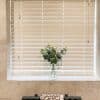 White horizontal blinds (Venetian), lowered and closed with side pull cords, filtering daylight; set in a tiled bathroom window recess with a glass vase of eucalyptus on the sill.