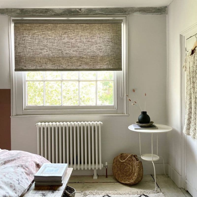 Woven roller blind lowered over upper half of a multi-pane window, filtering daylight; bedroom with white walls, radiator beneath, round side table with black vase and woven basket.