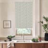 A floral-patterned roller blind covers a kitchen window, filtering daylight above a stainless sink; countertop holds a bowl of greens, soap, potted plants and a framed sketch.