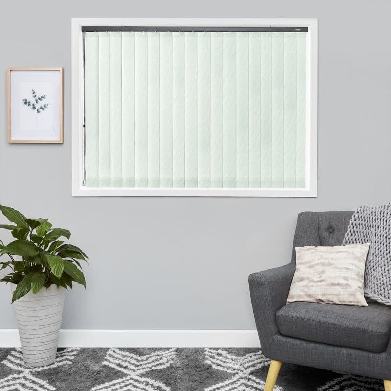 Vertical fabric blinds, pale mint textured slats hanging from a black headrail inside a white-framed window, closed and filtering daylight; on a grey living-room wall with plant, framed print and armchair.