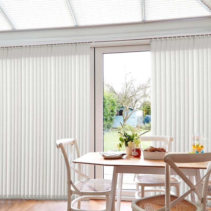 White floor-to-ceiling vertical blinds with narrow slats, parted at center; filtering soft daylight and revealing a glass patio door; a cozy dining area with wooden table, chairs, flowers, backyard view.