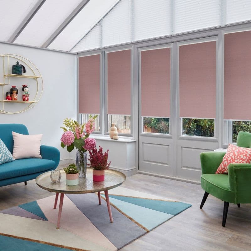 Pink roller blinds cover upper sections of tall conservatory windows and doors, partially lowered to filter light, in a sunroom with colorful seating, rug, and flowers on a coffee table.