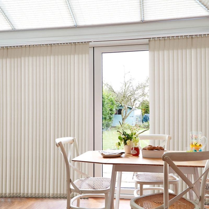 Beige vertical blinds, ceiling-mounted with pleated slats, partially parted to reveal a glass patio door, filtering soft daylight across a bright dining area with wooden table and chairs.
