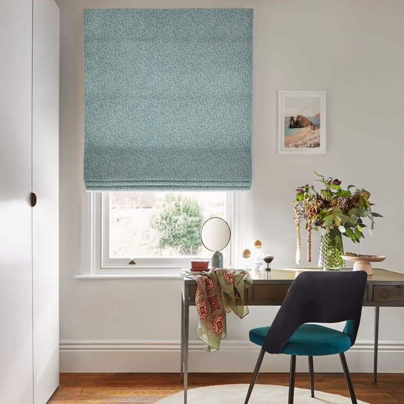 Teal patterned Roman blind, covering the upper window and partially lowered, filtering daylight through the lower sash; desk with mirror, vase of foliage and a chair in a bright neutral home office.