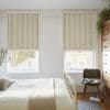 Beige-striped Roman blinds, lowered and partially folded, filtering soft daylight; bedroom with cream metal bed, wooden chest of drawers, green chair, plants and a view of neighboring brick facades.