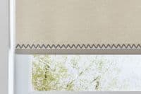 A beige fabric roller blind with a dark zigzag-stitched hem, partially lowered and filtering light, over a white window frame in a bright interior, revealing blurred green tree branches outside.