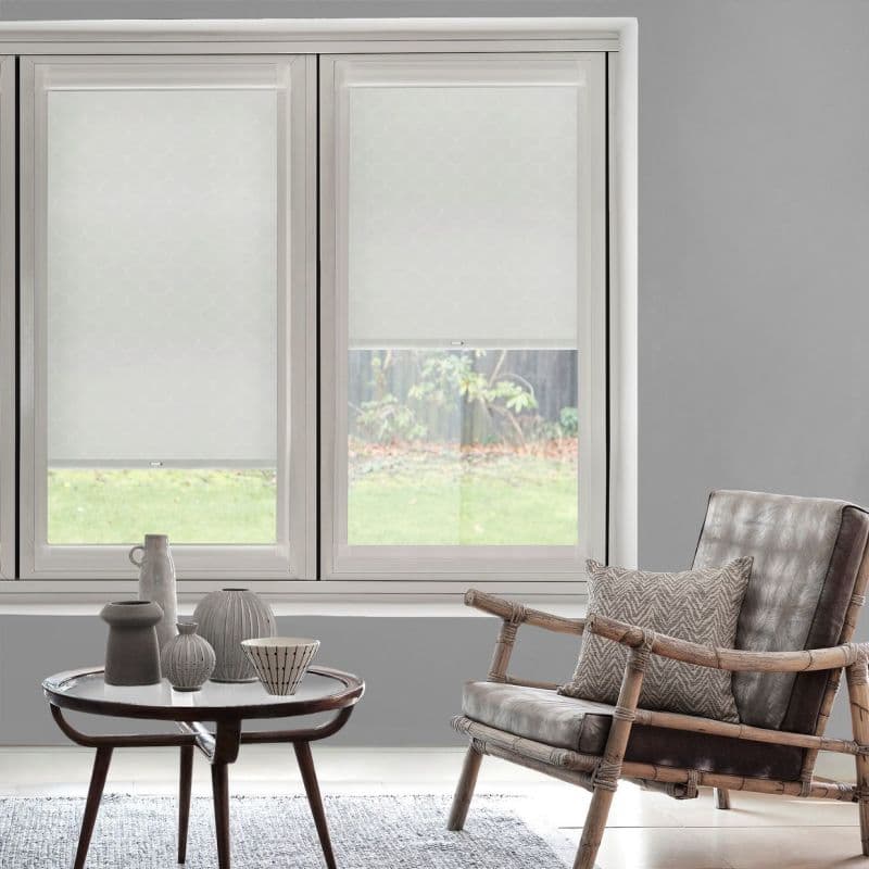 Two white patterned roller blinds, lowered across a double window with an opening on the right, filtering soft daylight; living room with gray walls, rattan armchair and coffee table.