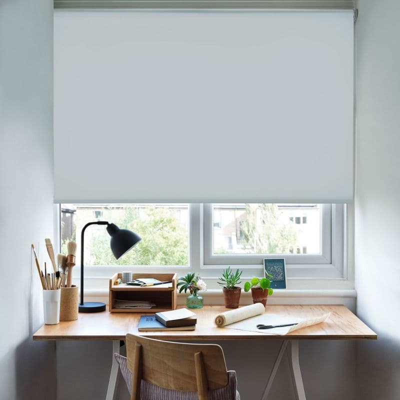 White fabric roller blind, lowered across the window, opaque, diffusing light and blocking most view. Small home desk beneath with lamp, brushes, notebooks, potted plants, and a wooden chair.
