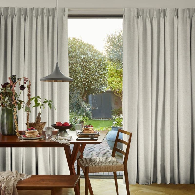 Floor-to-ceiling pleated cream curtains, slightly parted at center, filtering soft daylight. Dining table with pancakes and pendant lamp faces sunlit backyard with trees and a shed.