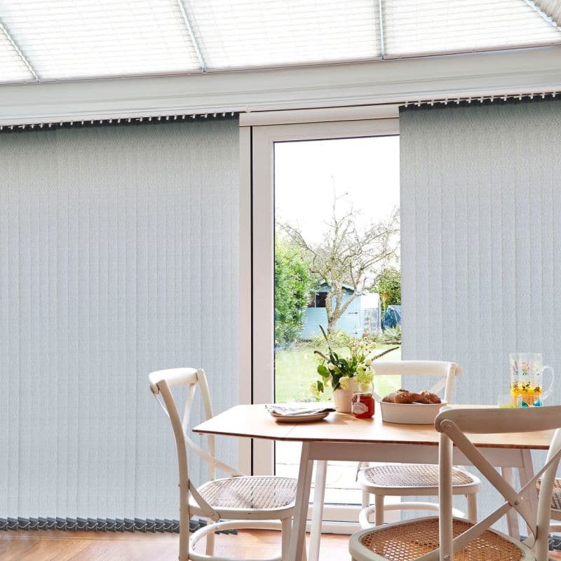 Vertical fabric blinds cover sliding doors — light-gray slats on a track, mostly closed with a central gap, filtering daylight into a dining nook with table, chairs and garden view.
