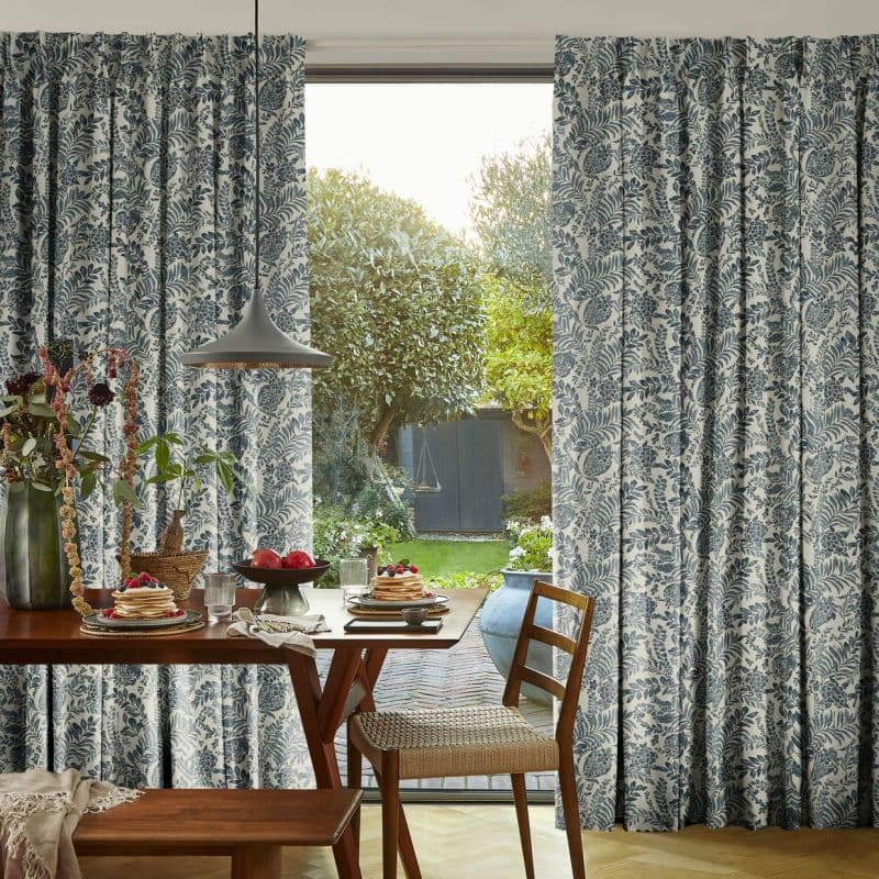 Floor-length pleated curtains with blue botanical print, mostly closed but parted to reveal a glass door; filtering sunlight into a dining area with wooden table set and sunlit garden beyond.