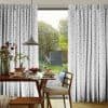 White pleated floor-length curtains with gray star pattern, parted at center to reveal a sunlit garden. They frame a dining table set for breakfast under a pendant lamp.