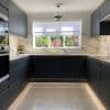 White roller blind with scalloped lower edge, lowered across a wide window, filtering daylight; modern U-shaped kitchen with dark cabinets, marble splashback, central sink and garden view with pink flowers.
