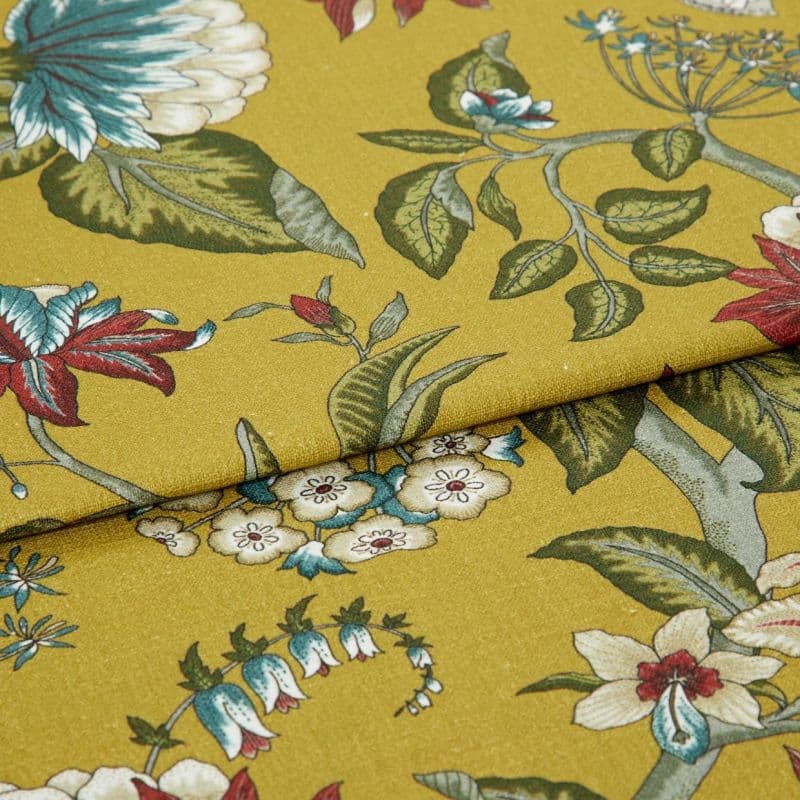 Mustard-yellow fabric with botanical green leaves and red-and-blue flowers, folded once with overlapping edge; close-up studio-lit view of the textile on a flat surface.