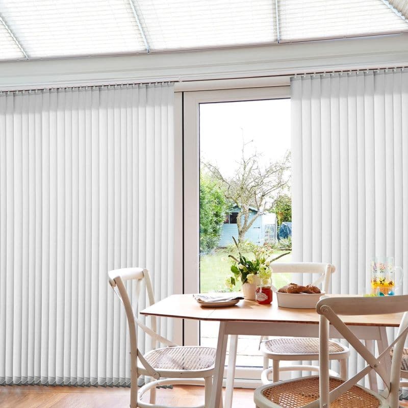 White vertical blinds with narrow fabric slats hang from a ceiling track, mostly closed but parted centrally to reveal a glass patio door; sunlight filters into a bright dining nook.