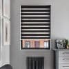 Black-and-gray zebra roller blind with alternating opaque and sheer horizontal stripes, lowered to filter light; in gray kitchen nook with espresso machine, radiator under window and brick building visible outside.