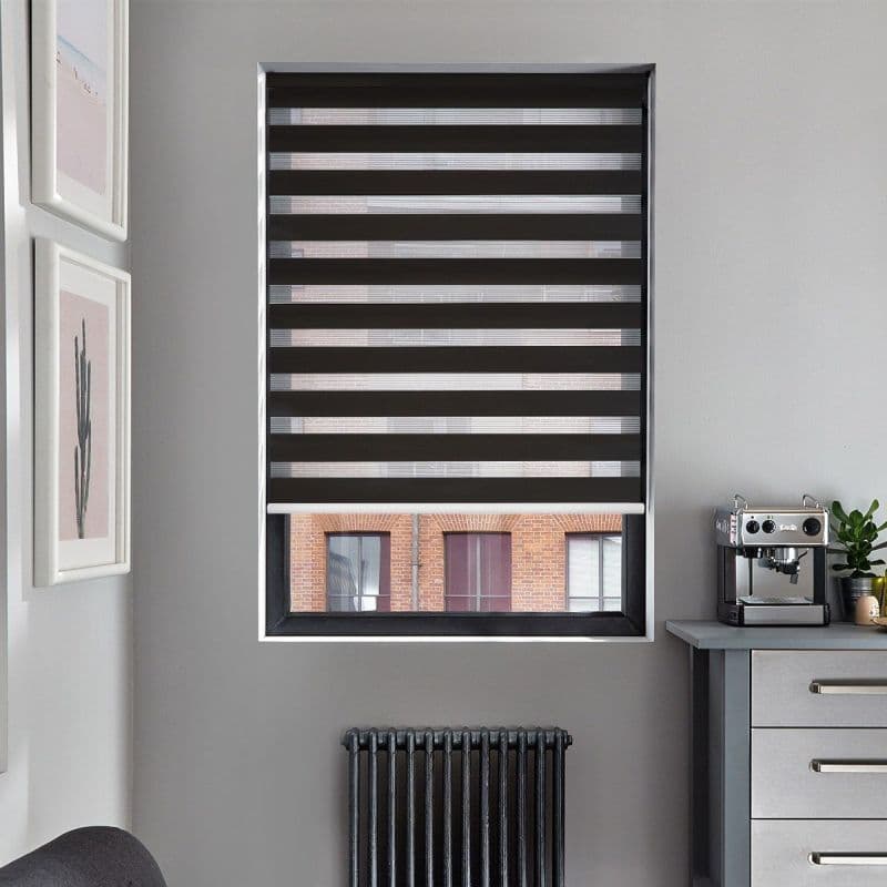 Black-and-gray zebra roller blind with alternating opaque and sheer horizontal stripes, lowered to filter light; in gray kitchen nook with espresso machine, radiator under window and brick building visible outside.