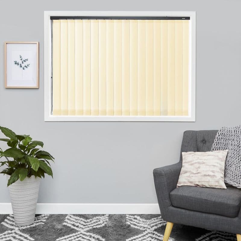 Cream vertical blinds across a central window—narrow slats on a black headrail—closed, softly filtering daylight, in a grey-walled living room with a potted plant, framed print, patterned rug, and grey armchair.