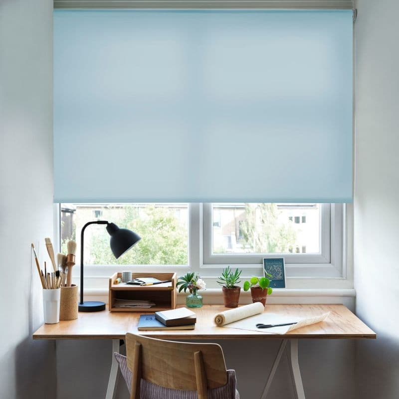 A pale blue roller blind, lowered across a double window, softly filtering daylight; a wooden desk underneath holds a lamp, brushes, notebooks and potted plants in a bright home workspace.
