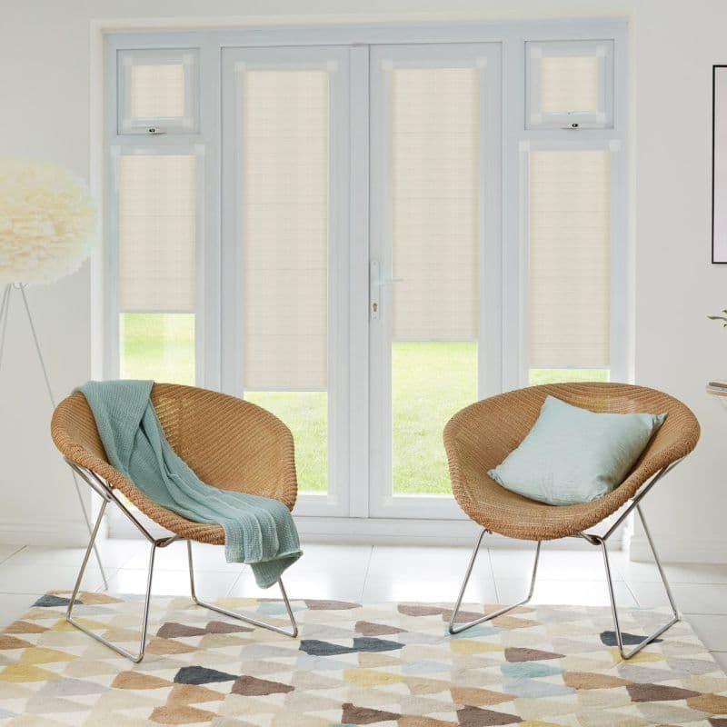 Beige roller blinds fitted inside white French doors and sidelights, lowered to partially filter daylight; bright living room with two wicker chairs, pastel throws, patterned rug and lawn view.