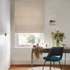 A beige diamond-patterned Roman blind, fully lowered with soft folds and filtering daylight; above a white sash window in a bright home office with desk, teal chair, vase of flowers.