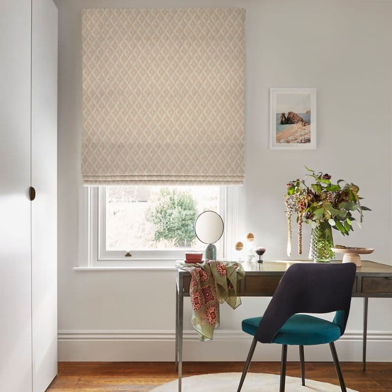 A beige diamond-patterned Roman blind, fully lowered with soft folds and filtering daylight; above a white sash window in a bright home office with desk, teal chair, vase of flowers.