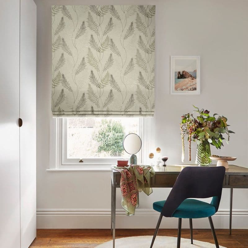 Roman blind with pale green fern pattern covering the window, lowered and filtering soft daylight; set above a desk with teal chair, vase of foliage, mirror and framed print.