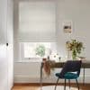 A light-gray textured Roman blind, lowered to just above the sill and softly filtering daylight, covers a window above a desk with mirror, vase of flowers and a teal chair.