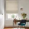 Beige Roman blind lowered partway with bottom folds, filtering daylight; above a small desk holding a mirror and vase of flowers, a teal chair sits in a neutral room.