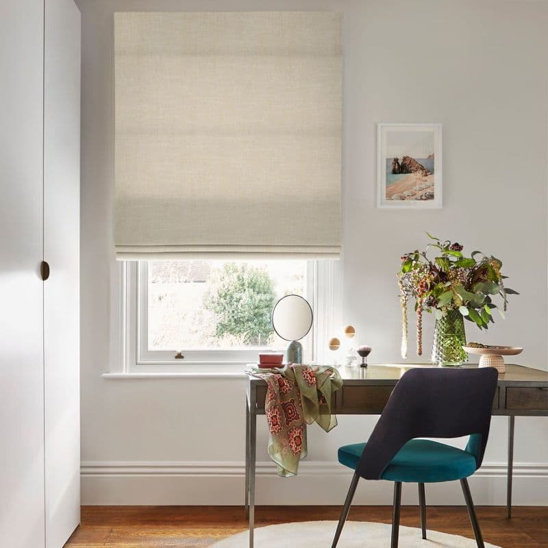 Beige Roman blind lowered partway with bottom folds, filtering daylight; above a small desk holding a mirror and vase of flowers, a teal chair sits in a neutral room.
