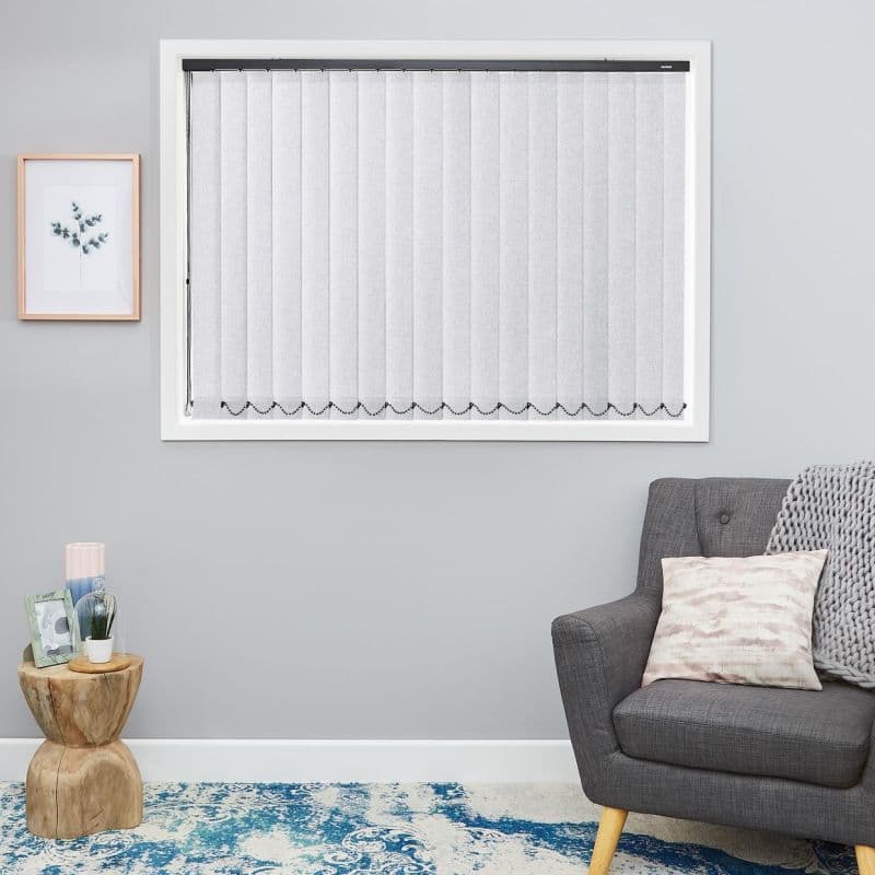 Vertical fabric blinds, closed and filtering light with a beaded bottom chain, cover a framed window; pale-gray walls, framed botanical print, gray armchair, knitted throw, wooden side table, blue rug.