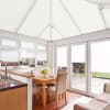 White pleated cellular blinds covering the pitched conservatory roof and window/door panels, closed and diffusing bright daylight; inside a light, neutral dining-kitchen conservatory with wooden table, chairs and patio view.