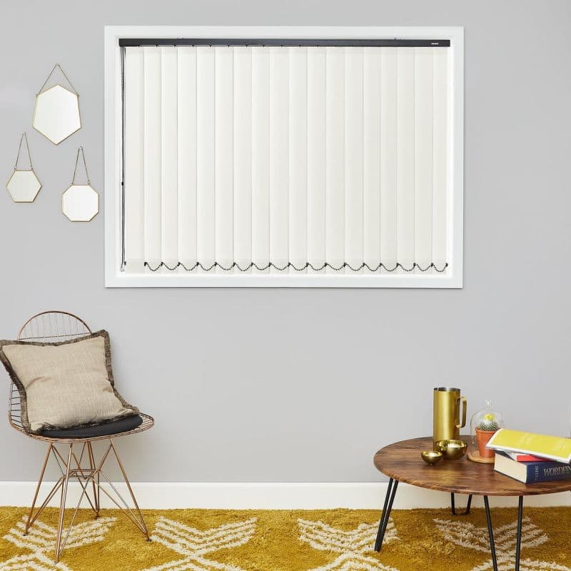 White vertical blinds (full‑length slats in a white frame) closed and filtering light; mounted on a grey living‑room wall above a wire chair, hexagonal mirrors, wooden coffee table, mustard rug.
WORDEN