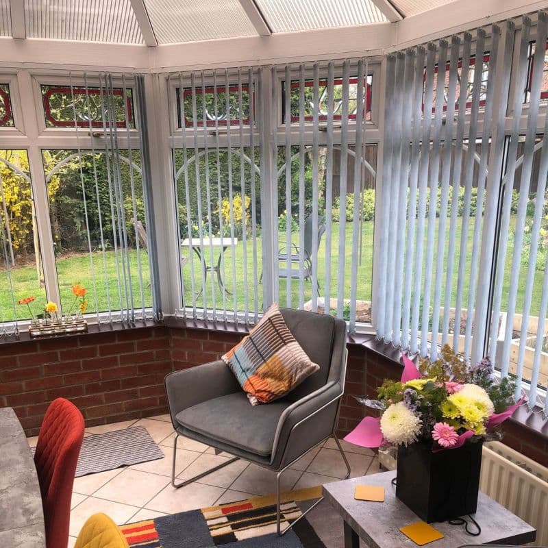 Light-grey vertical blinds cover the conservatory bay windows, ceiling-to-sill and slightly tilted to filter daylight; a tiled sunroom contains a grey armchair with patterned cushion and a bouquet, garden beyond.